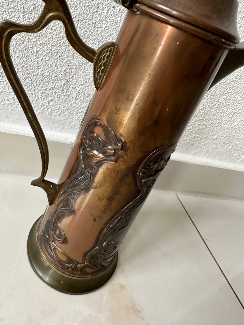May include: A copper pitcher with an ornate design of stylized flowers and leaves. The pitcher has a brass handle and a brass base.