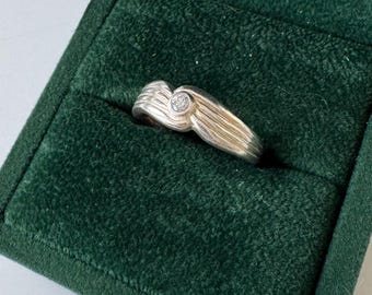 Puede incluir: Un anillo de plata con un solo diamante redondo y transparente engastado en el centro. El anillo está en una caja de anillo de terciopelo verde.