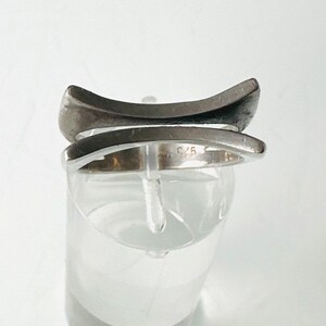 May include: A silver ring with a unique, modern design. The ring is made of two bands that intersect, creating a geometric shape. The ring is marked with the number "925", indicating that it is made of sterling silver.