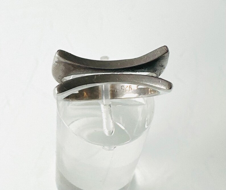 May include: A silver ring with a unique, modern design. The ring is made of two bands that intersect, creating a geometric shape. The ring is marked with the number "925", indicating that it is made of sterling silver.