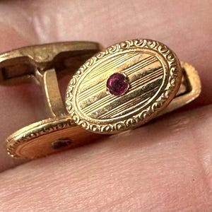 May include: A pair of gold cufflinks with an oval design. Each cufflink features a central, round, red gemstone and a patterned surface with etched lines. The edges are decorated with a detailed, ornate design.