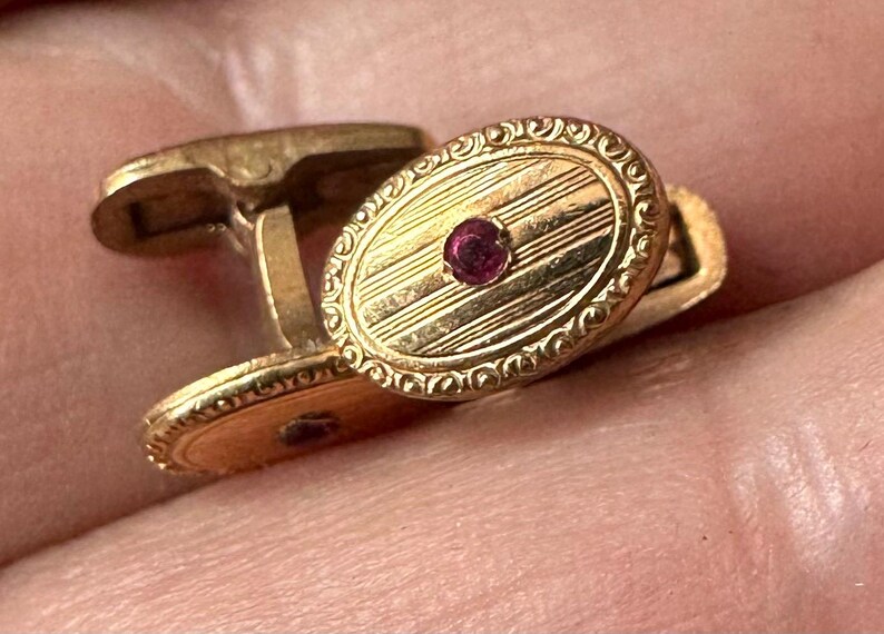 May include: A pair of gold cufflinks with an oval design. Each cufflink features a central, round, red gemstone and a patterned surface with etched lines. The edges are decorated with a detailed, ornate design.