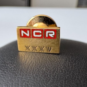 Könnte beinhalten: Ein goldfarbener NCR-Ansteckpin mit den Buchstaben "NCR" in roten Kästchen. Unter den Buchstaben stehen die römischen Ziffern "XXXXV". Der Pin hat eine runde Rückseite und ist wahrscheinlich ein Vintage-Sammlerstück oder ein Firmenartikel.