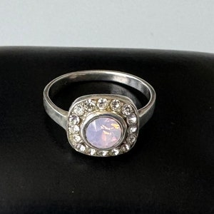 Puede incluir: Un anillo de plata con un engaste cuadrado que presenta un ópalo rosa rodeado de diamantes de imitación transparentes.
