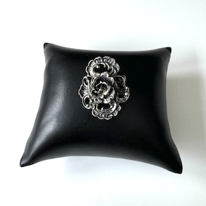 May include: A silver filigree brooch in the shape of a rose. The brooch is on a black velvet cushion.