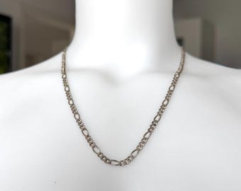 May include: Silver-toned Figaro chain necklace displayed on a white mannequin. The necklace features a pattern of three small circular links followed by one elongated oval link. The chain is a classic jewelry piece.