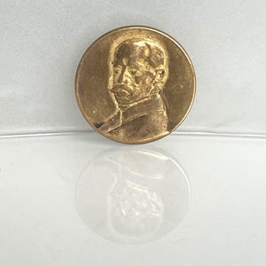 Könnte beinhalten: Eine kreisförmige, goldfarbene Metallmedaille mit einem erhabenen Reliefporträt eines Mannes mit Schnurrbart im Profil. Die Medaille hat eine glatte, polierte Oberfläche mit einem ausgeprägten metallischen Glanz, der das Licht reflektiert.