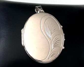 May include: An oval-shaped silver-toned metal locket pendant featuring a smooth, polished surface on the left side and a decorative engraved pattern of three stylized leaf-like shapes on the right side, complete with a small top bail for hanging.