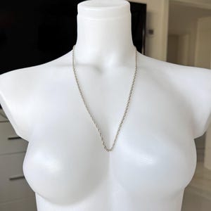 Peut inclure: Un collier chaîne de couleur argentée présenté sur un mannequin blanc. Le collier est de conception simple et délicate, suspendu en forme de V. Le mannequin est dans une pièce bien éclairée, avec un arrière-plan flou.