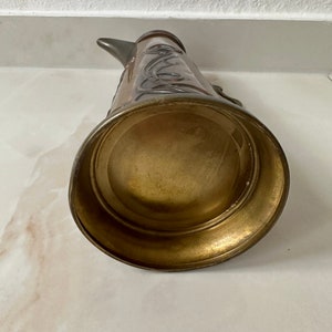 May include: A vintage copper and brass pitcher with an ornate design. The pitcher has a wide base and a narrow spout.