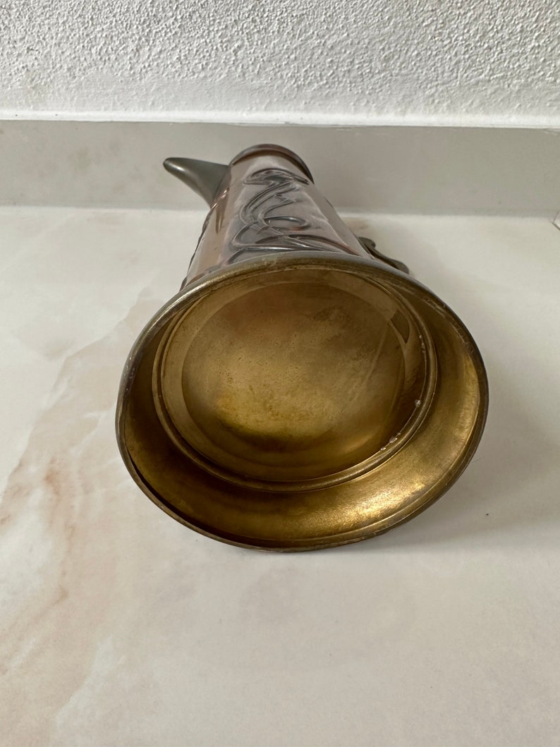 May include: A vintage copper and brass pitcher with an ornate design. The pitcher has a wide base and a narrow spout.