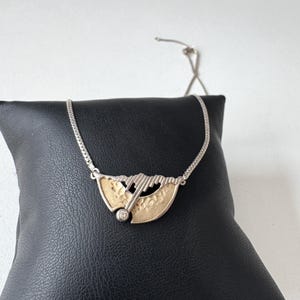 Puede incluir: Un collar de plata con un diseño de colgante único. El colgante presenta un elemento dorado en forma de abanico con una superficie texturizada, detalles plateados y una pequeña gema transparente. El collar se muestra sobre una superficie negra.