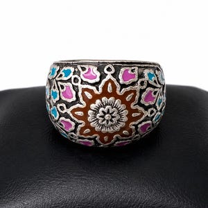 Könnte beinhalten: Ein breiter silberfarbener Ring mit einem markanten braunen, sternförmigen Motiv in der Mitte, umgeben von komplizierten Filigranmustern, die mit leuchtend rosa und hellblauen Emaille-Akzenten gefüllt sind und eine detaillierte Oberfläche bilden.