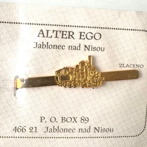 Könnte beinhalten: Goldener Krawattenclip mit einem detaillierten Lokomotivdesign. Der Krawattenclip befindet sich auf einer weißen Karte mit dem Text "ALTER EGO Jablonec nad Nisou ZLACENO P. O. BOX 89 466 21 Jablonec nad Nisou".