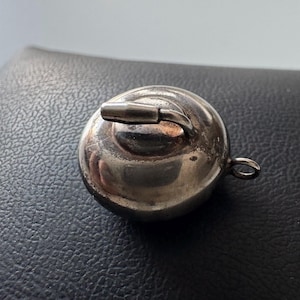 May include: A small, silver-colored pendant in the shape of a curling stone. The pendant has a handle on top and a small loop for a chain. The surface is reflective, and the pendant is resting on a textured black surface.