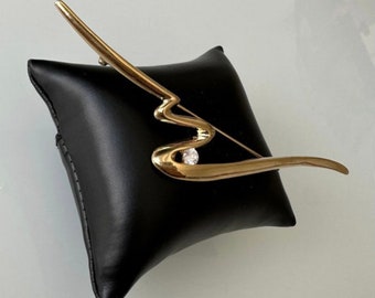 May include: A gold-toned brooch with a wavy, abstract design and a single clear rhinestone in the center.