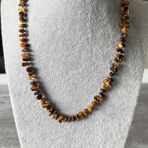 May include: A brown and gold tiger's eye stone necklace on a gray mannequin. The necklace is made of small, irregular shaped beads.