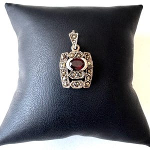 Könnte beinhalten: Ein silberner Anhänger mit rechteckigem Design, der einen großen ovalen, tiefroten Edelstein in der Mitte aufweist. Der Anhänger ist mit kleineren, funkelnden Steinen verziert und wird auf einer schwarzen Oberfläche präsentiert.