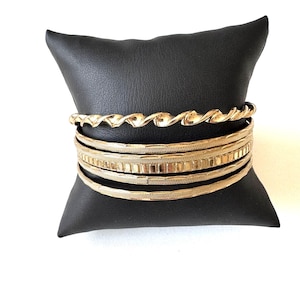 Könnte beinhalten: Eine Sammlung goldfarbener Armreifen, präsentiert auf einem schwarzen Kissen. Die Armreifen weisen unterschiedliche Designs auf, darunter ein gedrehter Seil-Stil und strukturierte Bänder. Der Schmuck ist vor weißem Hintergrund platziert, was den metallischen Glanz hervorhebt.