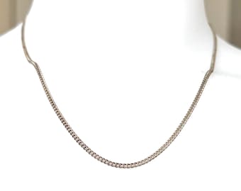 May include: A silver-colored curb chain necklace displayed on a white mannequin. The chain has small, interlocking links and is designed to be worn around the neck. The necklace is a simple, elegant piece of jewelry.