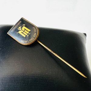 Peut inclure: Une épingle dorée avec un design en forme de bouclier noir et or. Le bouclier porte le texte "MÜNCHEN BOWLING DM 1968" et une image stylisée d'un quille de bowling.