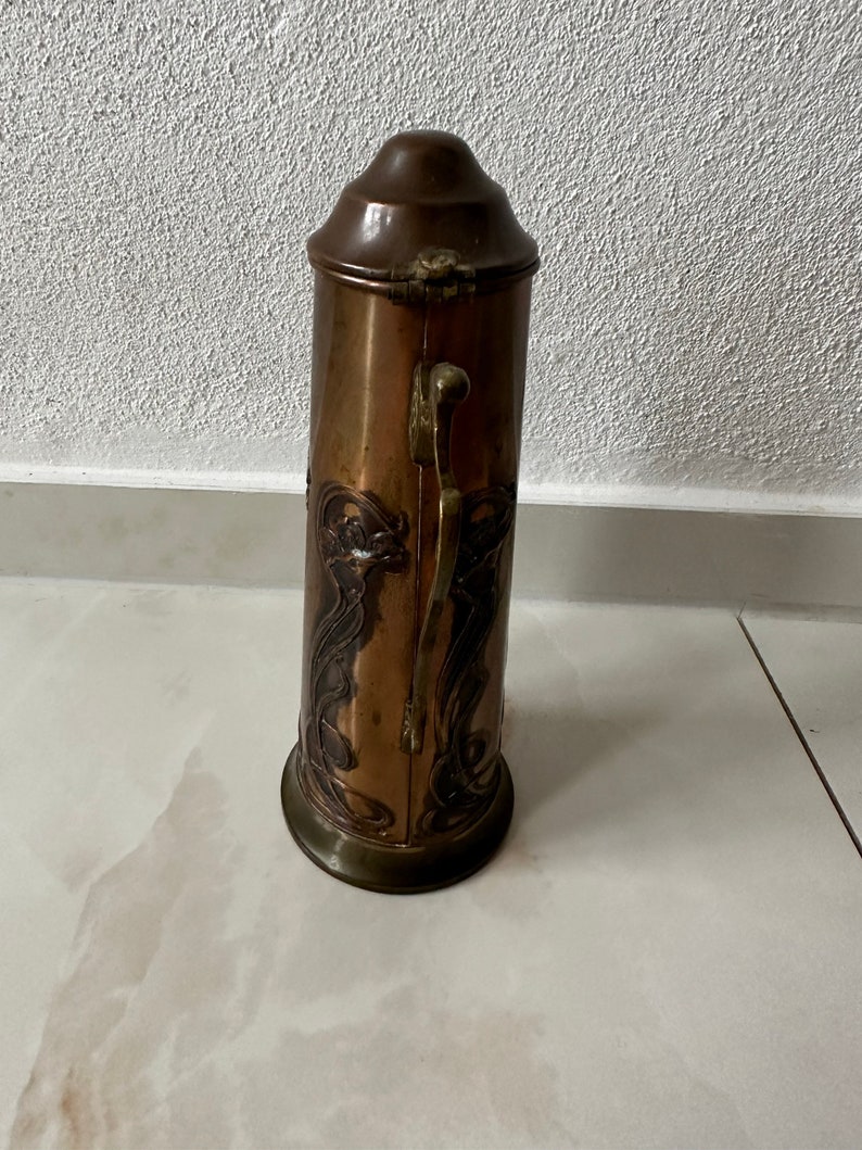May include: A copper and brass vintage boot scraper with an ornate Art Nouveau design. The scraper has a hinged lid and a handle on the side.