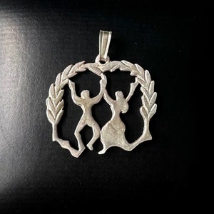 May include: A silver pendant featuring a stylized design of two figures dancing within a wreath of leaves.