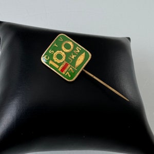 May include: A green and gold pin with the text "CSTV 100 JKM" and a red stripe. The pin is on a black surface.