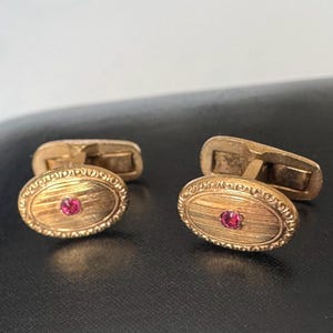 May include: A pair of gold-toned cufflinks with an oval design. Each cufflink features a small, round, red gemstone in the center and a textured, engraved border. The cufflinks are displayed on a dark surface.
