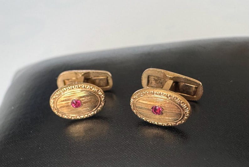 May include: A pair of gold-toned cufflinks with an oval design. Each cufflink features a small, round, red gemstone in the center and a textured, engraved border. The cufflinks are displayed on a dark surface.