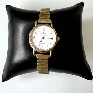 Op de afbeelding: Een goudkleurig Lorus quartz horloge met een ronde witte wijzerplaat, zwarte Arabische cijfers en dunne zwarte wijzers. Het horloge heeft een flexibele, rekbare metalen schakelband in goudkleur die om een zwart vierkant kussen is gewikkeld.