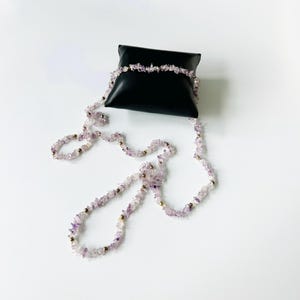 May include: A long, delicate necklace made of small, irregular amethyst stones and gold beads. The necklace is displayed on a black velvet cushion.