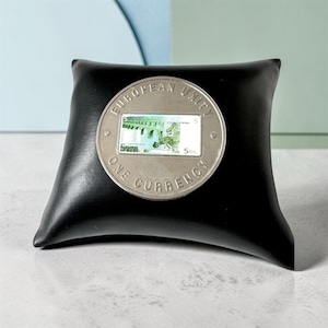 May include: A circular silver-toned coin featuring the text EUROPEAN UNITY ONE CURRENCY and a central rectangular inlay depicting a green architectural bridge design with the number 5 and the word EURO printed on the coin surface.