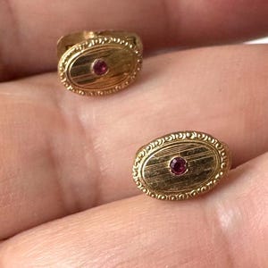 May include: Two gold-colored cufflinks with an oval design. Each cufflink features a small, round, red gemstone in the center. The edges of the cufflinks have a detailed, ornate pattern. The cufflinks are likely vintage or antique.
