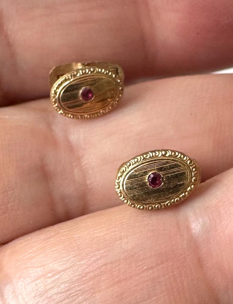 May include: Two gold-colored cufflinks with an oval design. Each cufflink features a small, round, red gemstone in the center. The edges of the cufflinks have a detailed, ornate pattern. The cufflinks are likely vintage or antique.