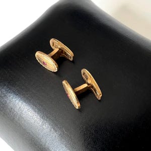 May include: A pair of gold-toned cufflinks with an oval design and a small red stone in the center. The cufflinks are displayed on a black surface, showcasing their polished finish and intricate details. The jewelry is a classic accessory.