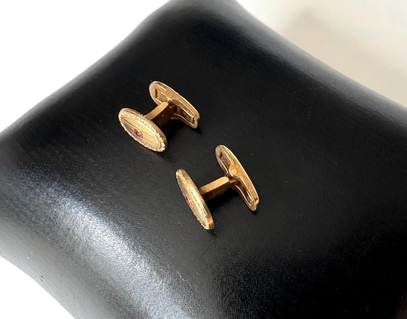 May include: A pair of gold-toned cufflinks with an oval design and a small red stone in the center. The cufflinks are displayed on a black surface, showcasing their polished finish and intricate details. The jewelry is a classic accessory.