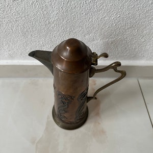 May include: A copper pitcher with an ornate design and a hinged lid. The pitcher has a handle and a spout.
