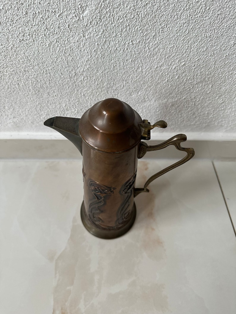 May include: A copper pitcher with an ornate design and a hinged lid. The pitcher has a handle and a spout.