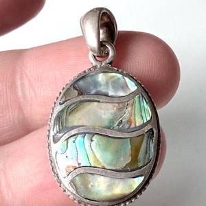 Op de afbeelding: Ovale hanger met iriserende abalone schelpinleg, met tinten blauw, groen en zilver. De hanger heeft een zilverkleurig metalen frame met een decoratieve rand en een lus voor een ketting.
