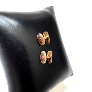 May include: A pair of gold-toned cufflinks with an oval design and a small red gemstone in the center. The cufflinks are displayed on a black leather surface, showcasing their elegant design and craftsmanship.