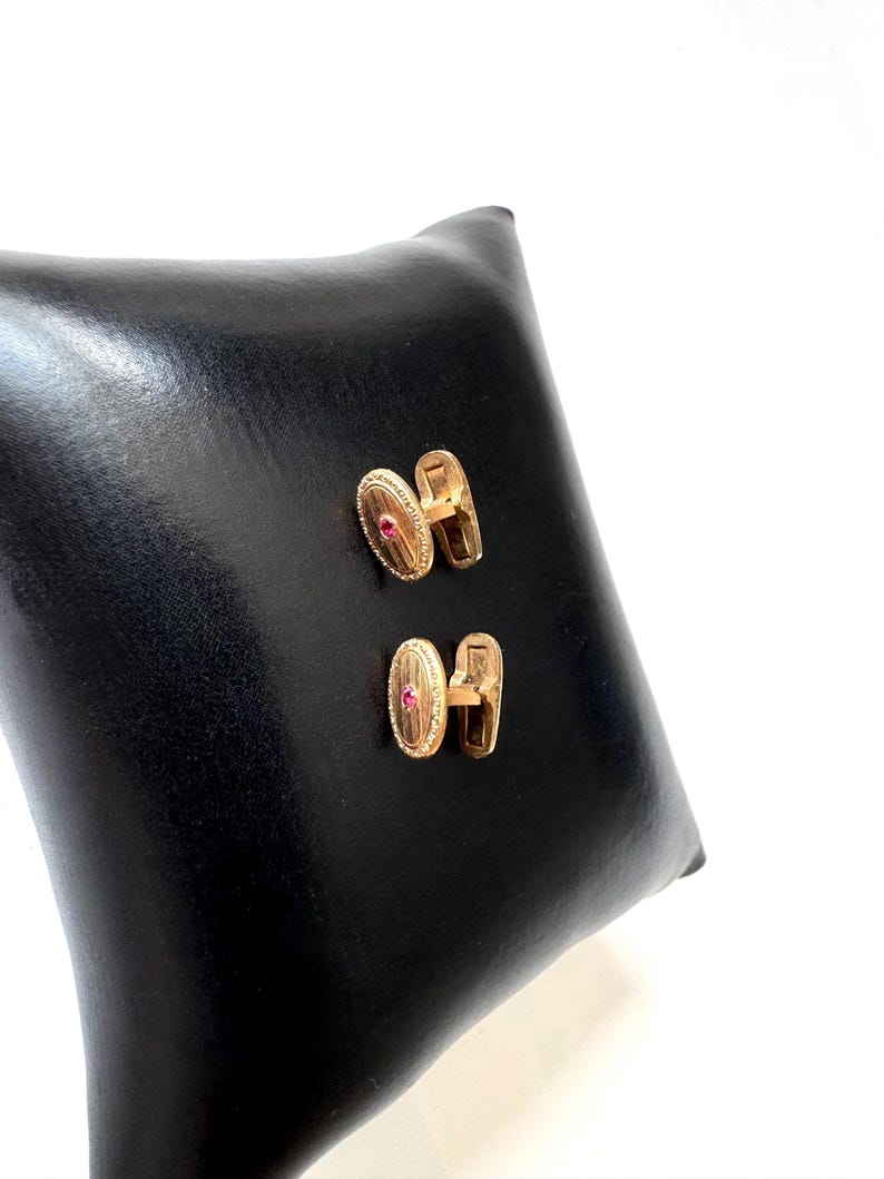 May include: A pair of gold-toned cufflinks with an oval design and a small red gemstone in the center. The cufflinks are displayed on a black leather surface, showcasing their elegant design and craftsmanship.