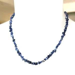 May include: A necklace composed of numerous small, irregular, tumbled blue stone chips. The stones vary in shades of deep navy, royal blue, and lighter sky blue, strung together to form a continuous strand that rests in a circular shape.