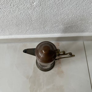 May include: A copper and brass teapot with a lid and a handle. The teapot is on a white surface.