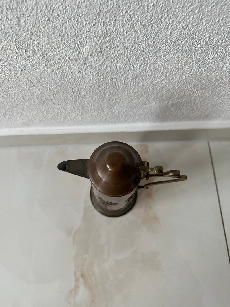 May include: A copper and brass teapot with a lid and a handle. The teapot is on a white surface.