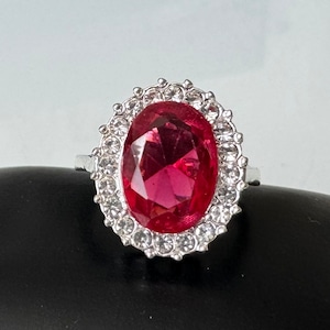May include: A silver ring featuring a large, oval-shaped, pink gemstone surrounded by smaller, clear gemstones.