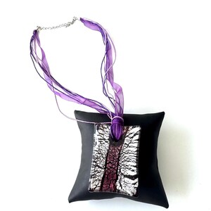 May include: A rectangular pendant necklace with a silver chain and purple ribbon. The pendant features a black and white pattern with a central purple stripe. The necklace is displayed on a black pillow.