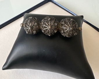 May include: A silver filigree bracelet featuring three spherical, openwork beads. The beads are connected by small, round links. The bracelet is displayed on a black leatherette pillow.