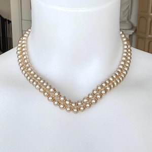 May include: A double-strand pearl necklace in a soft, creamy hue. The necklace is displayed on a white mannequin, highlighting its elegant design. The pearls are uniformly sized and closely strung, offering a classic and timeless appearance.