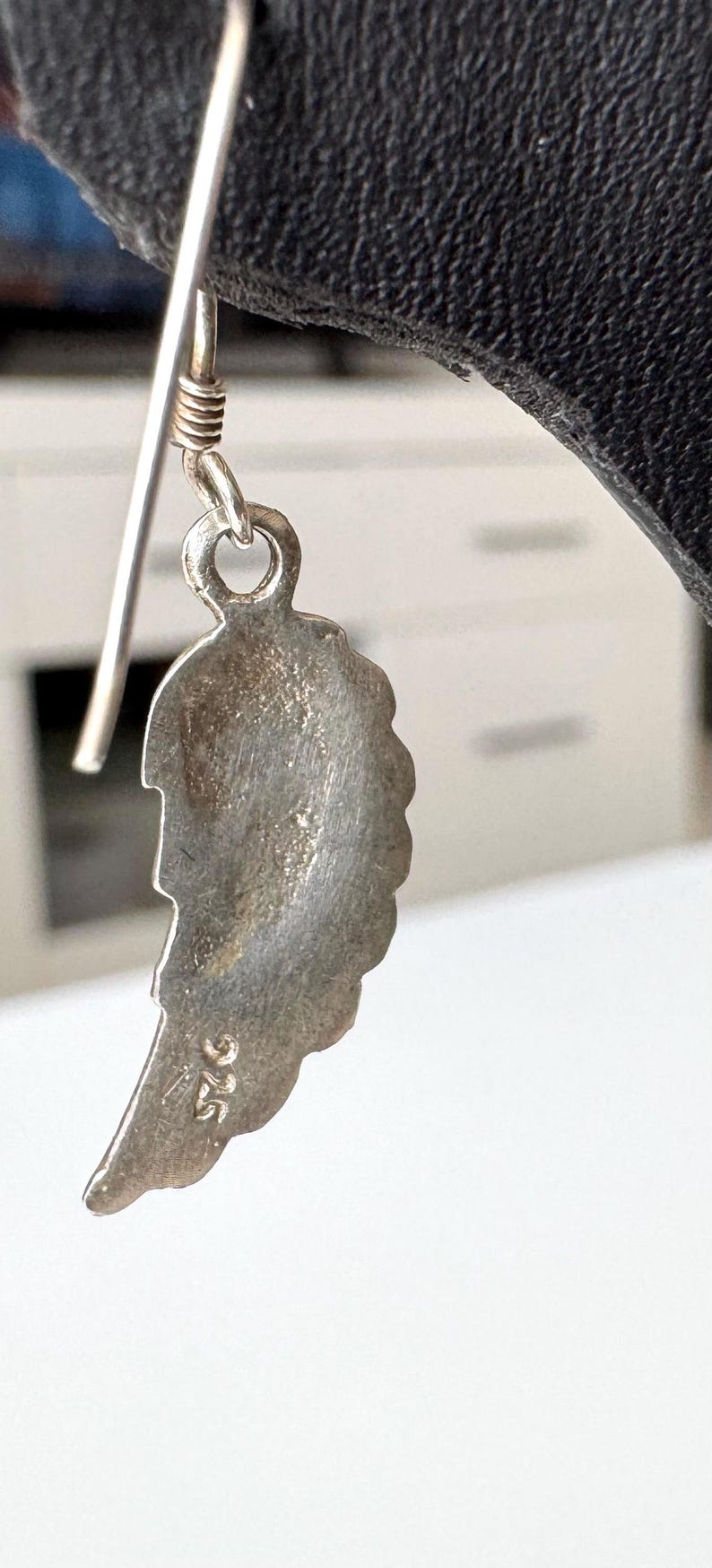 Pu&ograve; includere: Primo piano di un orecchino d'argento con un design a forma di piuma. L'orecchino ha un gancio e un piccolo anello. La piuma ha una superficie strutturata e il numero "925" &egrave; stampato in basso.